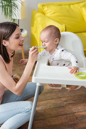 happy woman feeding little son sitting in baby chair with closed eyes