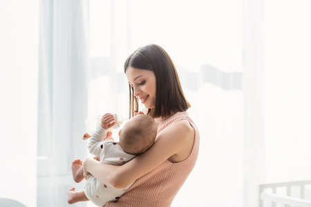 happy brunette woman feeding toddler from baby bottle while standing near white curtainの写真素材