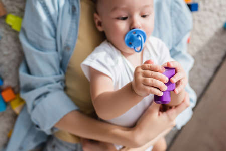 blurred little boy with pacifier playing with building block near motherの写真素材
