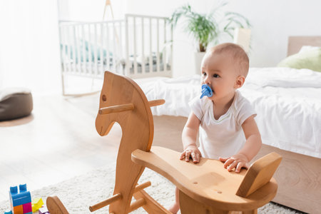 toddler child with pacifier standing near rocking horse, blurred bed and cribの写真素材