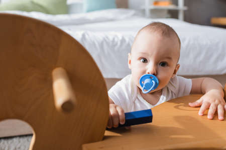 baby with pacifier holding toy building block while looking at camera near blurred rocking horseの写真素材