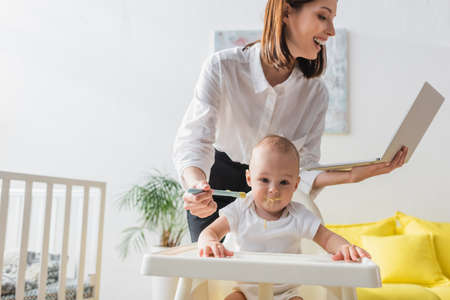 happy woman looking at laptop while feeding toddler son sitting in baby chairの写真素材