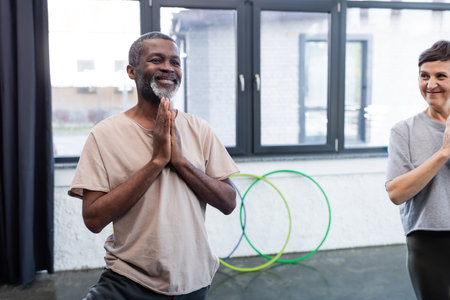 Smiling african american man practicing yoga near senior friend in sports centerの写真素材