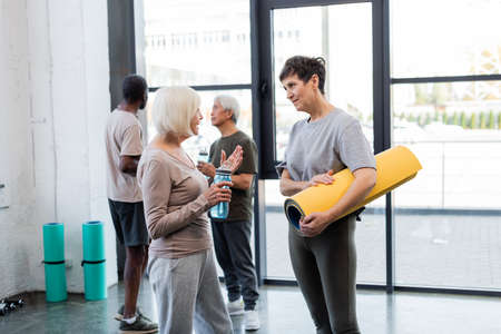 Senior women with fitness mat and sports bottle talking in gym
