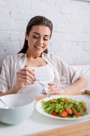 cheerful pregnant woman holding fork near tasty meal on trayの写真素材