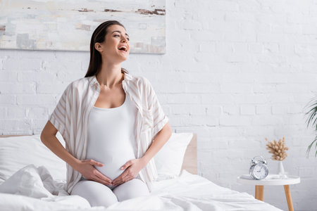 happy pregnant woman laughing and touching belly in bedroomの写真素材