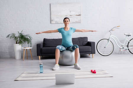 cheerful pregnant woman in sportswear exercising on fitness ball near laptop in living roomの写真素材