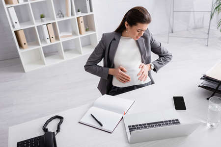 high angle view of displeased pregnant businesswoman touching belly while feeling cramp in office