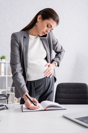 pregnant businesswoman writing in notebook and talking on cellphoneの写真素材