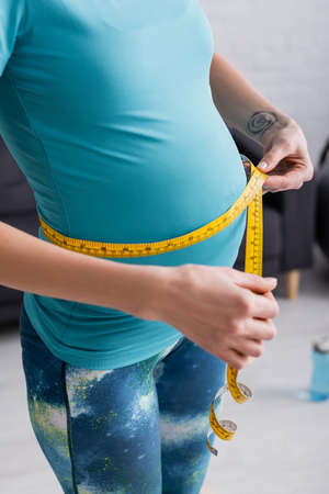 cropped view of tattooed pregnant sportswoman measuring belly at homeの写真素材