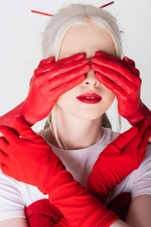 Female hands in red paint covering eyes of albino model isolated on whiteの写真素材