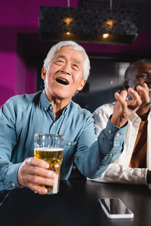 amazed asian man with glass of beer watching championship in sports bar near senior multiethnic...