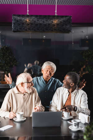 amazed asian man showing wow gesture near senior interracial friends pointing at laptop in cafeの写真素材