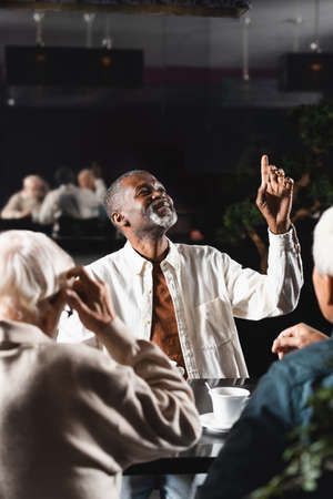 smiling african american man looking up and pointing with finger near blurred senior friends in cafeの写真素材
