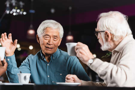 senior asian man gesturing while drinking coffee and talking with multiethnic friendsの写真素材