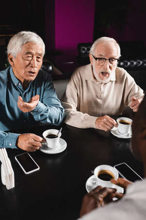 senior interracial men gesturing while talking to blurred african american friend in cafeの写真素材
