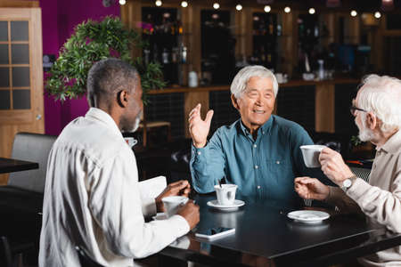 smiling asian man gesturing while talking with senior multiethnic friends in cafeの写真素材