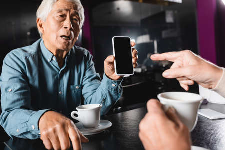 senior asian man holding smartphone with blank screen near blurred friend pointing with fingerの写真素材