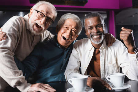 smiling african american man showing rejoice gesture near laughing senior friends in cafeの写真素材