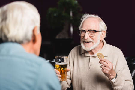 smiling elderly man holding glass of beer and chips near blurred friend in pubの写真素材