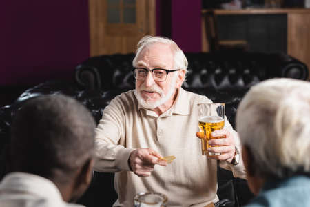 cheerful senior man holding chips and beer while talking to blurred interracial friends in pubの写真素材