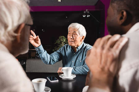 senior asian man looking away and pointing with finger near blurred interracial friendsの写真素材