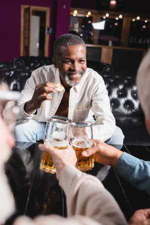 happy senior african american man holding chips while clinking glasses of beer with blurred friendsの写真素材