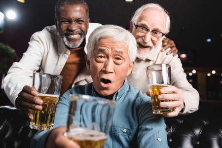 cheerful senior multicultural friends looking at camera while holding glasses of beer in pubの写真素材