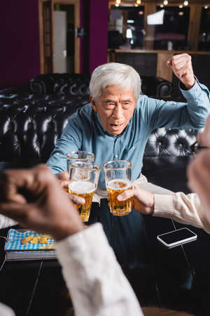 excited senior asian man showing win gesture while clinking beer glasses with blurred interracial friendsの写真素材