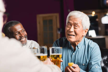 happy asian man holding beer and chips near blurred multiethnic friends in pubの写真素材