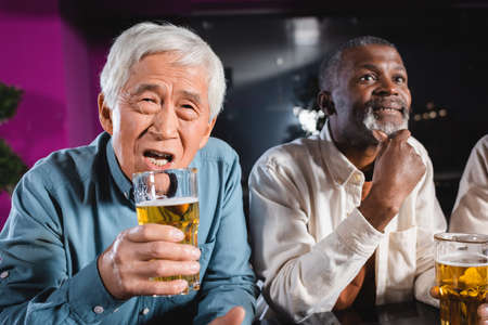 worried asian man holding glass of beer while watching football with interracial friends in pubの写真素材