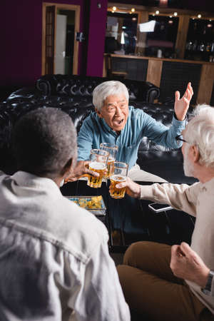 thrilled asian man gesturing while clinking glasses of beer with senior interracial friends in pubの写真素材