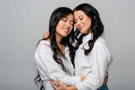 joyful asian mother and young daughter in white shirts embracing isolated on greyの写真素材