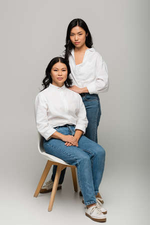 full length of asian mother sitting on chair near daughter in blue jeans and white shirt on greyの写真素材