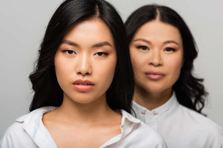 asian young adult daughter near blurred mother standing behind isolated on greyの写真素材