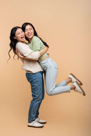 full length of cheerful asian mother lifting daughter on beigeの写真素材
