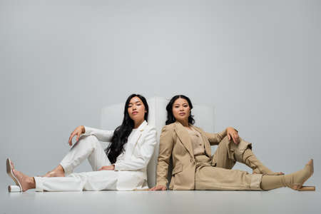 full length of trendy asian young adult woman sitting with brunette mother near cube on greyの写真素材