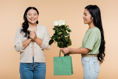 happy asian daughter giving paper bag and flowers to smiling mother isolated on beigeの写真素材