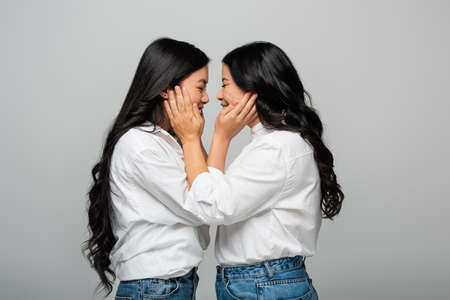 side view of happy asian mother and young adult daughter touching cheeks of each other isolated on greyの写真素材