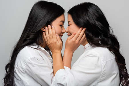 side view of cheerful asian mother and young adult daughter touching cheeks of each other isolated on greyの写真素材