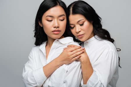 brunette asian mother and young adult daughter hugging each other and holding hands isolated on greyの写真素材