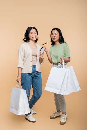 full length of cheerful asian mother and daughter with shopping bags and credit cards on beigeの写真素材