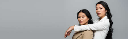 young asian woman hugging brunette mother in beige suit isolated on gray, bannerの写真素材