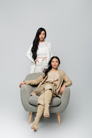 full length of asian woman sitting in armchair near trendy young daughter standing with hand in pocket on greyの写真素材