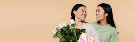 cheerful asian woman looking at mother with flowers and greeting card isolated on beige, bannerの写真素材