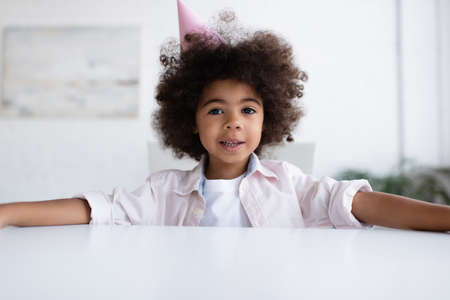 joyful african american birthday girl in party cap smiling at camera at homeの写真素材