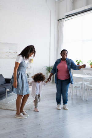 cheerful african american women holding hands and dancing with toddler girl at homeの写真素材