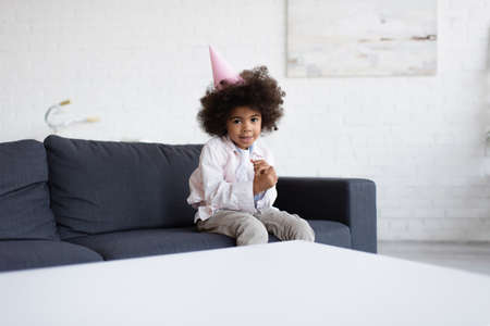 african american birthday girl in party cap sitting on couch near blurred table and looking at cameraの写真素材