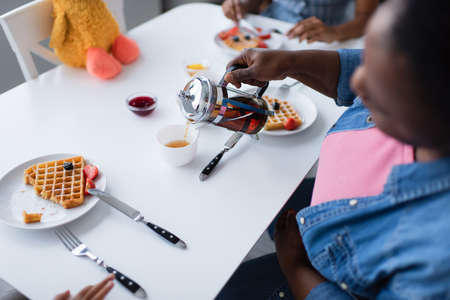 high angle view of blurred african american woman pouring tea near tasty waffles with berries during breakfast with familyの写真素材