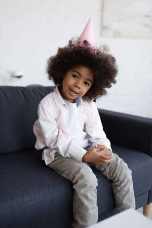 curly african american child in party cap looking at camera while sitting on couchの写真素材